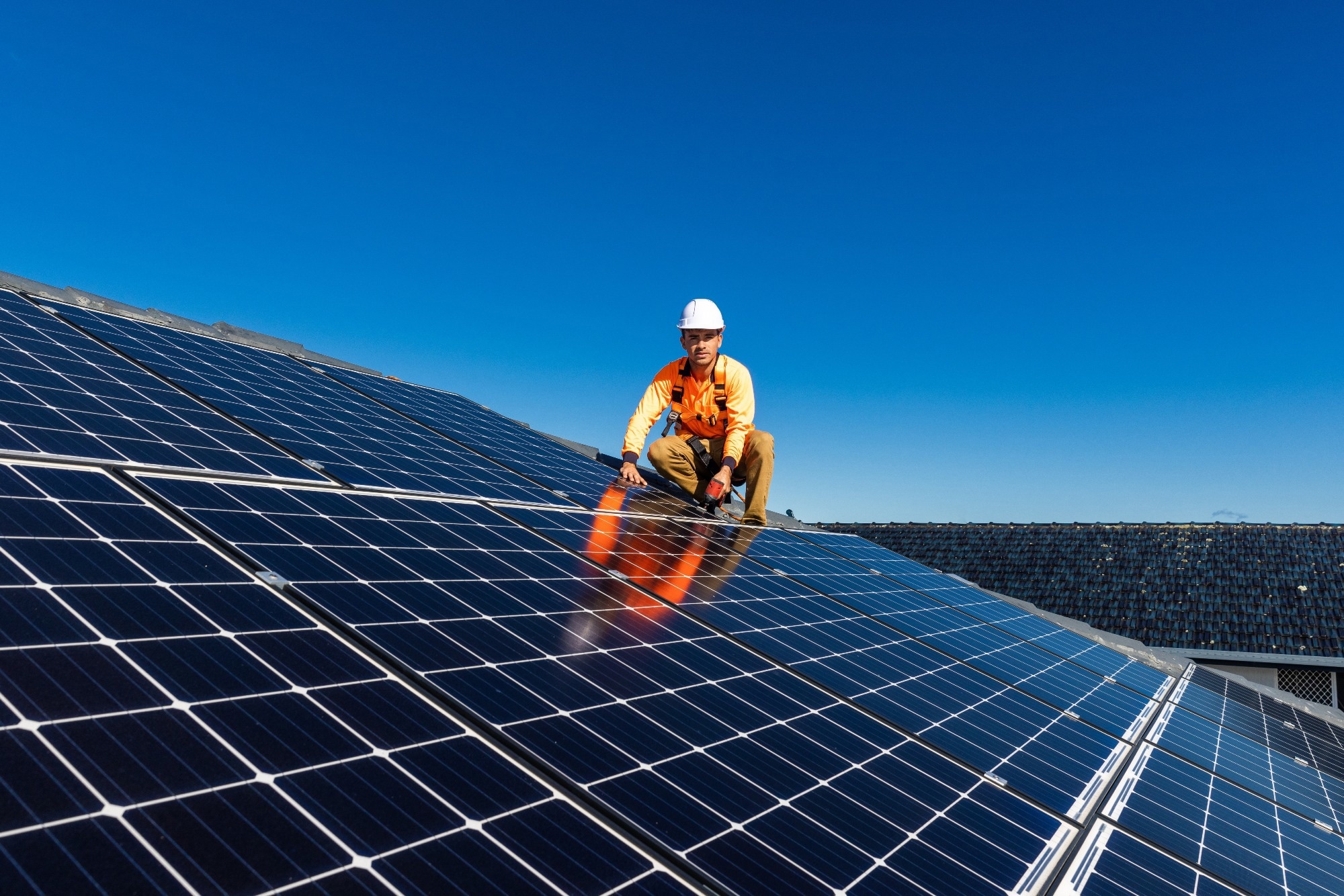 Solar panel technician with drill installing solar panels on house roof on a sunny day.