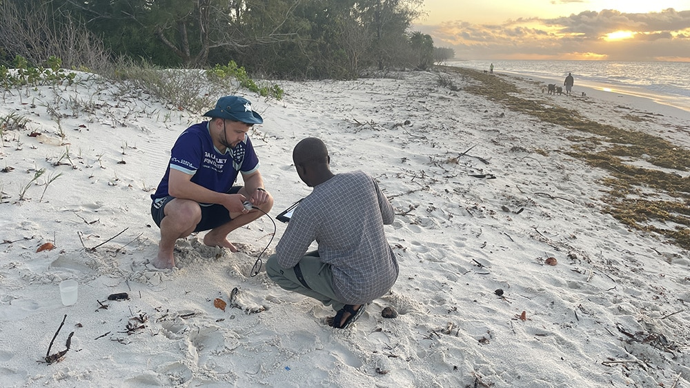 Fred Baggs and Ishmael Munda from LOC crouching on a sandy beach, smiling at the camera and holding a WET150 Senso.