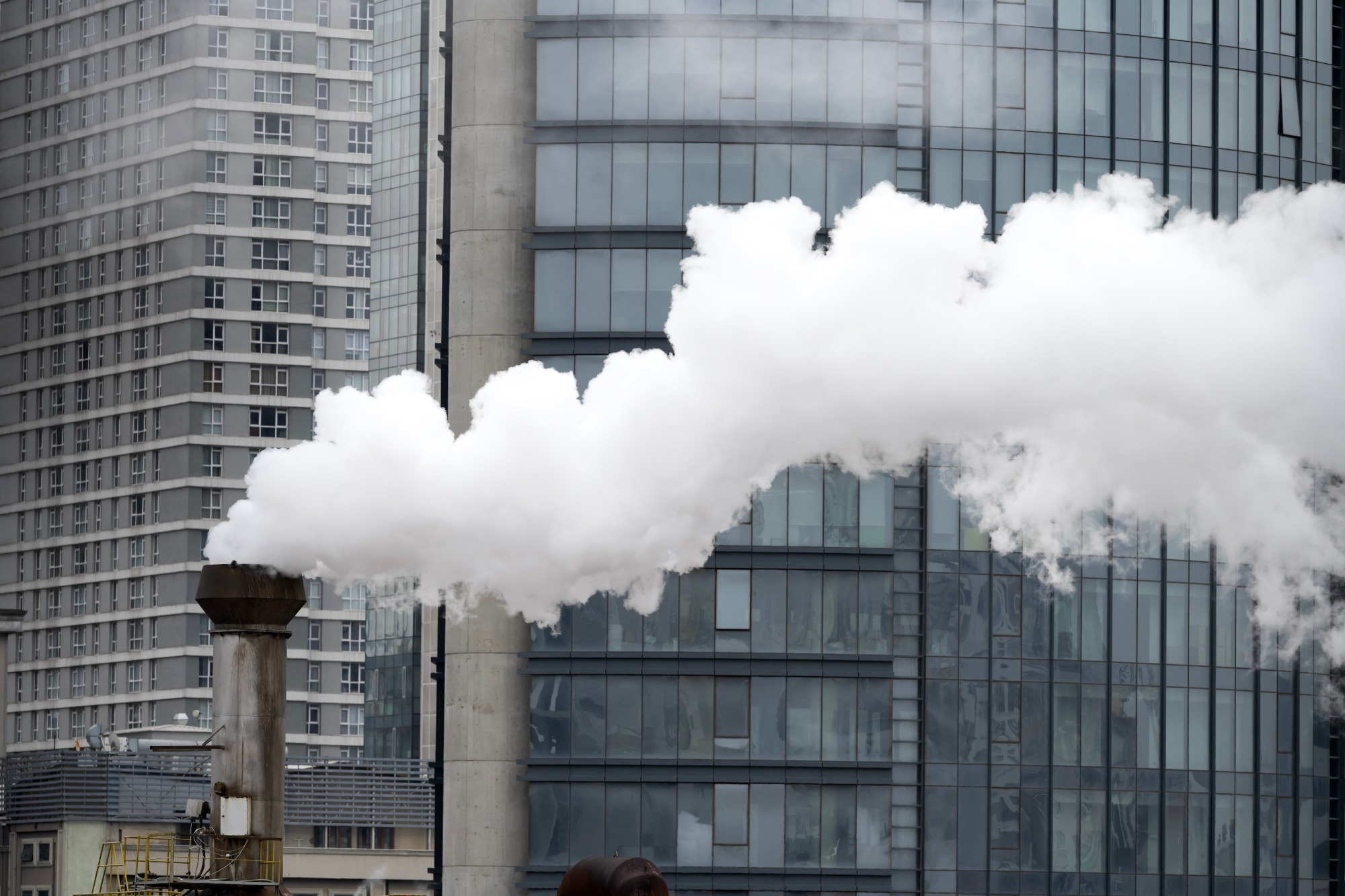A chimney emitting white smoke against a background of skyscrapers.