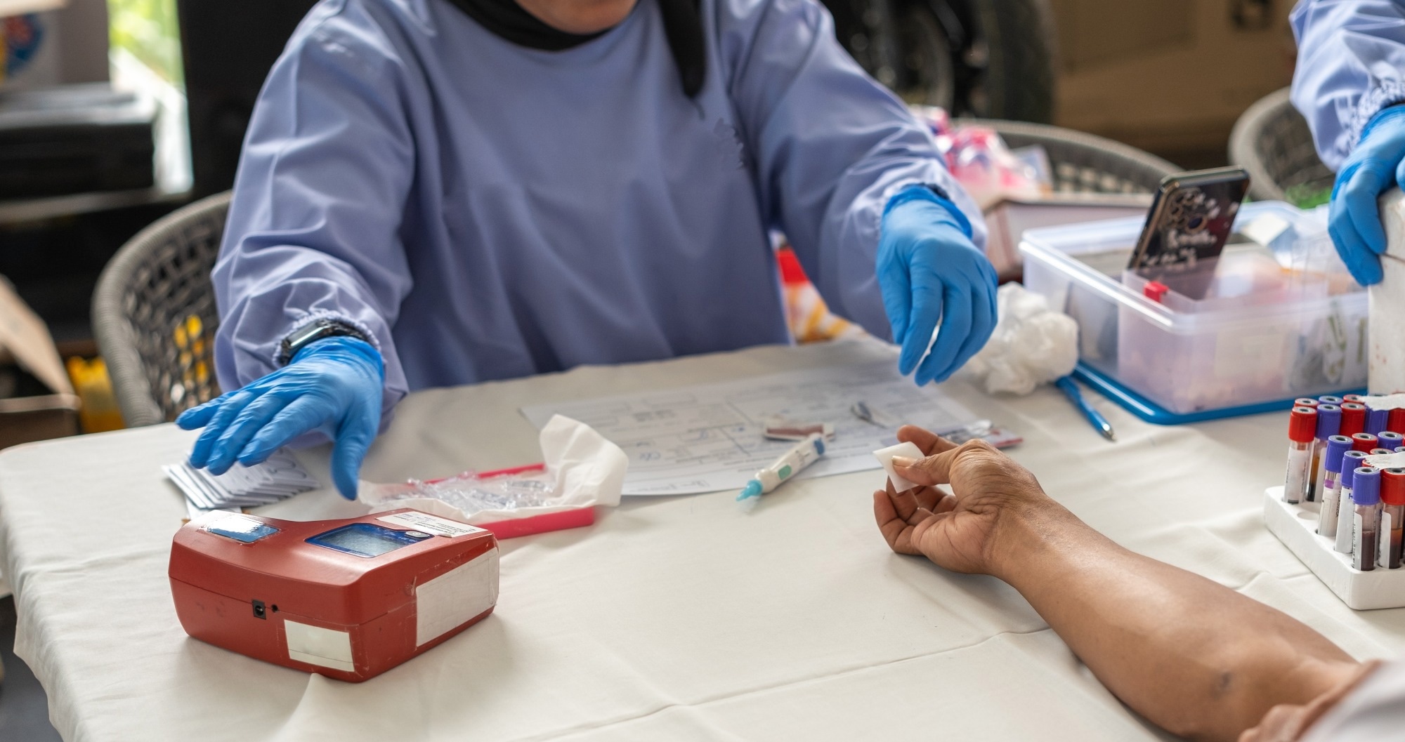 A healthcare worker wearing gloves prepares to collect a blood sample from a donor during a medical procedure. The table is equipped with medical tools and blood sample tubes