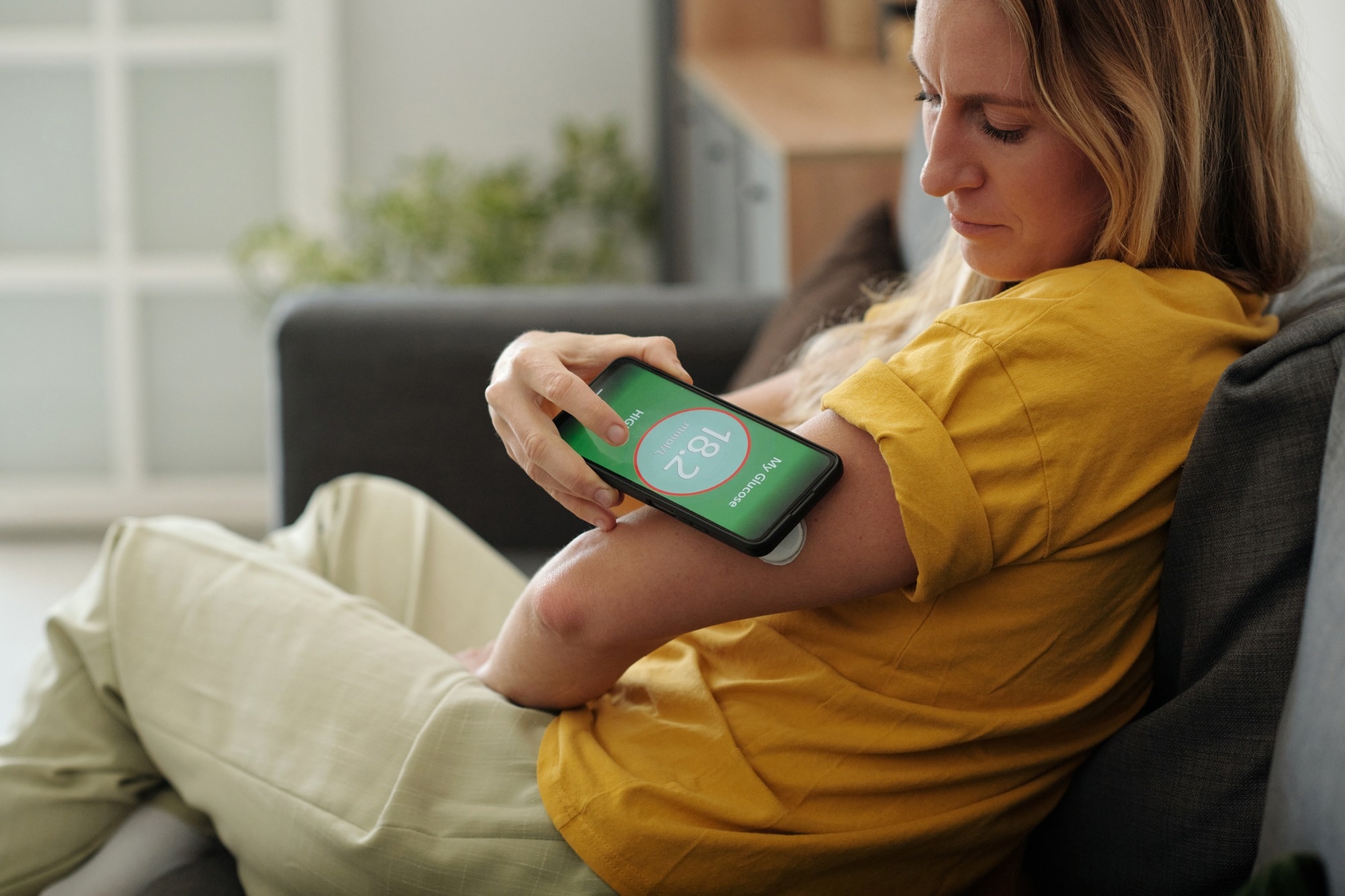 Side view closeup of woman checking glucose levels on phone with monitoring device on arm while sitting on couch.