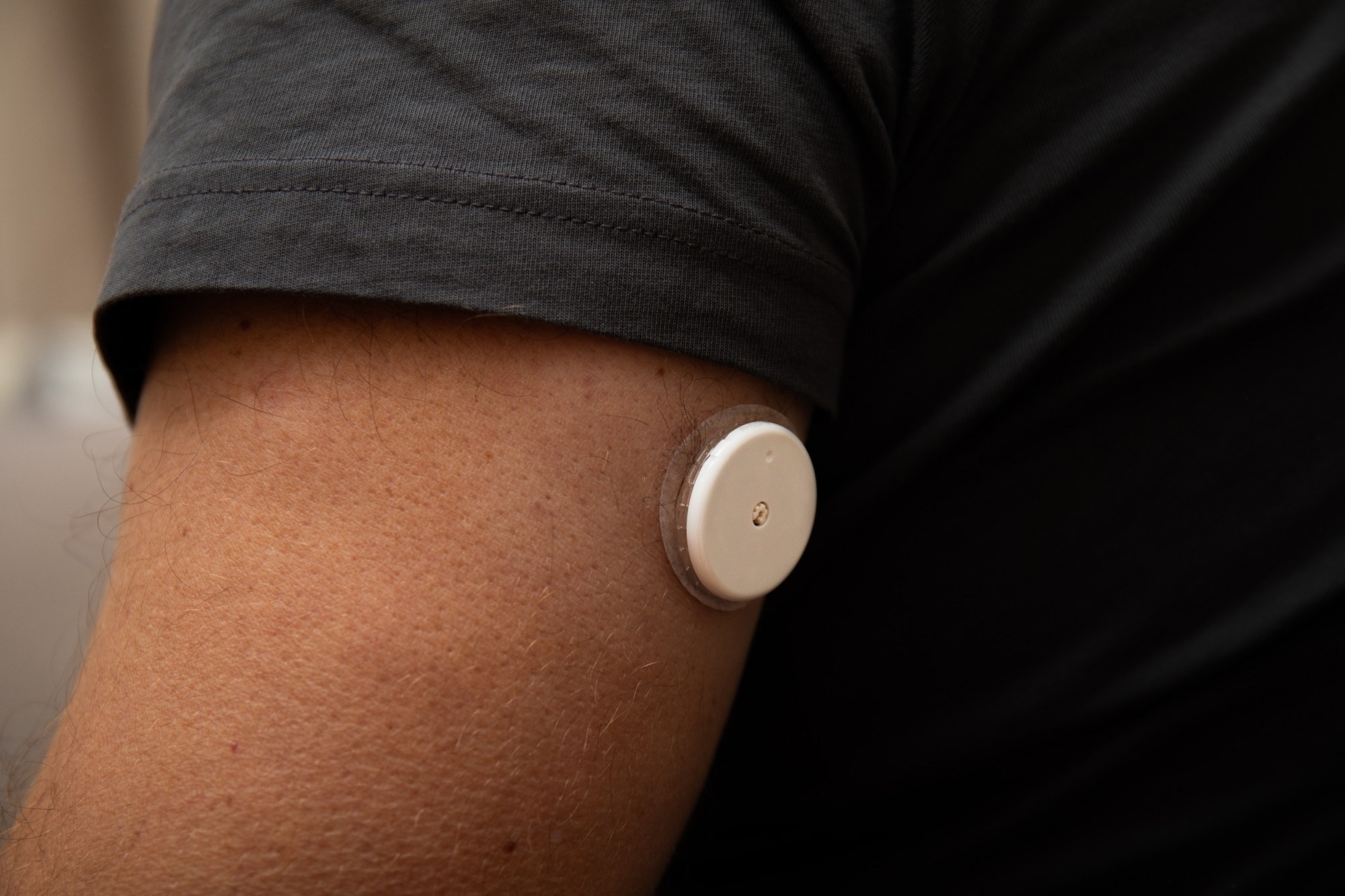 Close-up of continuous glucose monitoring device attached to upper arm, man in black t-shirt, neutral indoor background