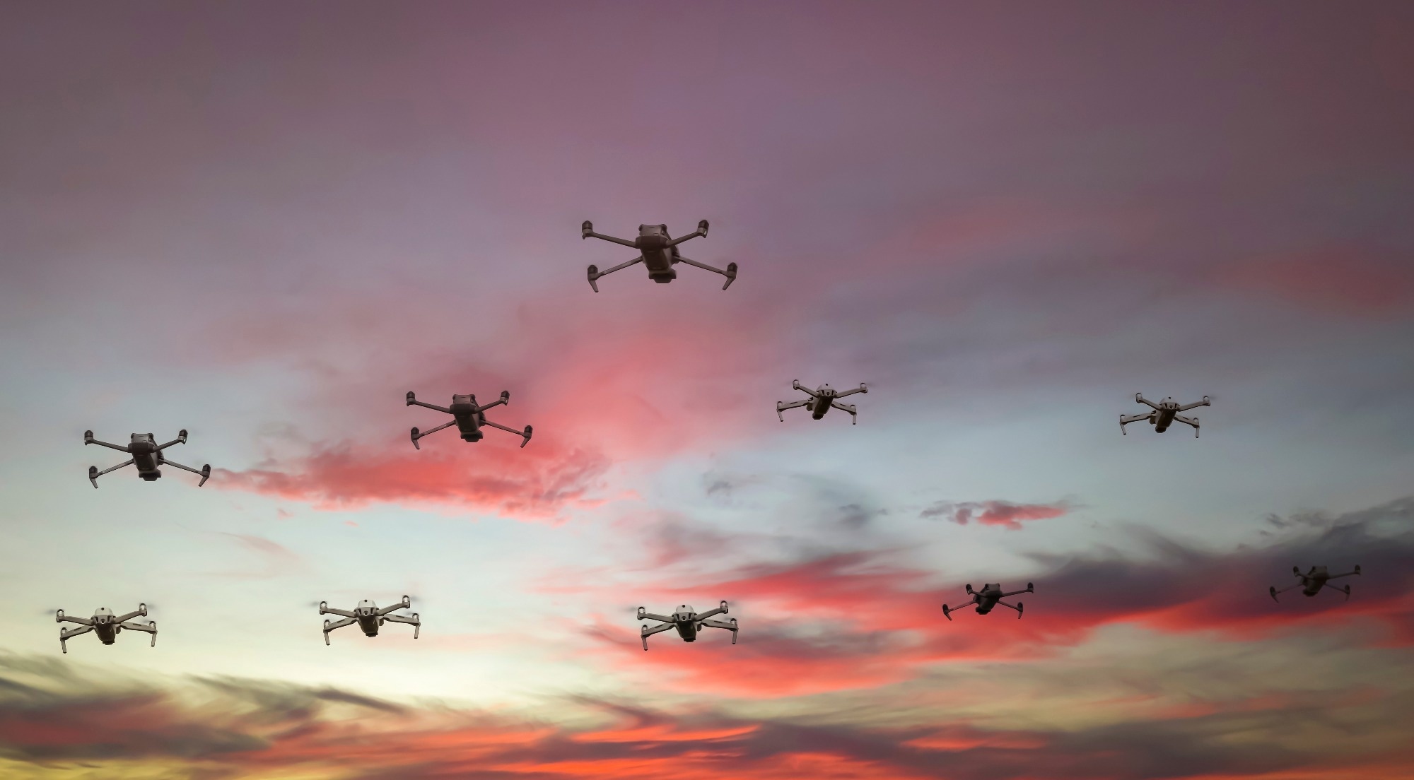 A group of drones fly in a sunset sky.