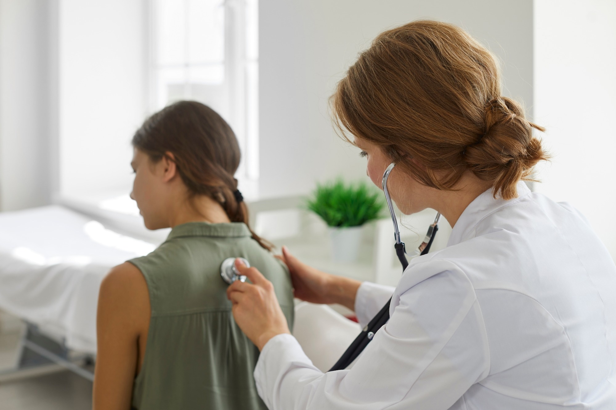 Female doctor in a white coat examining the lungs of a teenage girl.