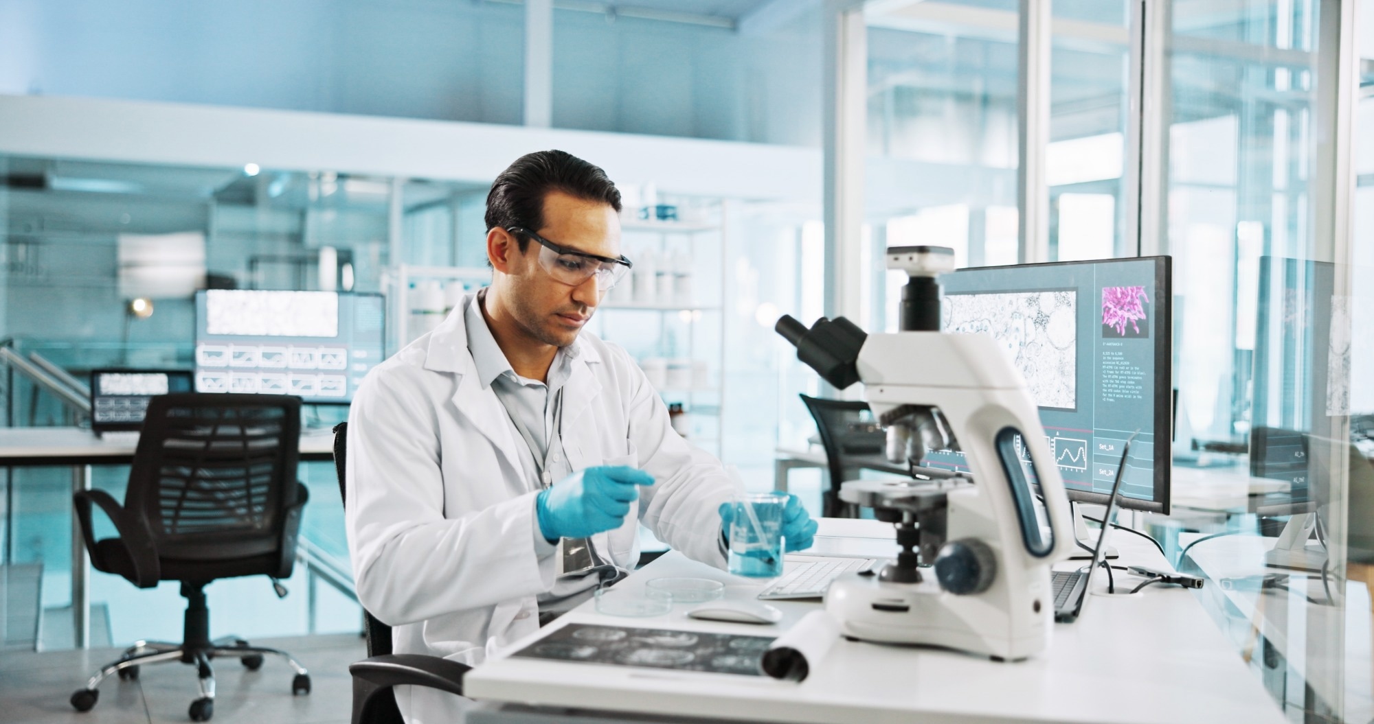 A scientist wearing goggles and a lab coat prepares a sample to examine under the microscope.
