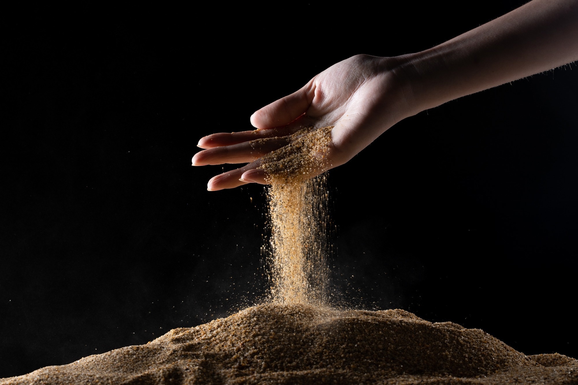 A hand against a black background pouring sand onto a surface.