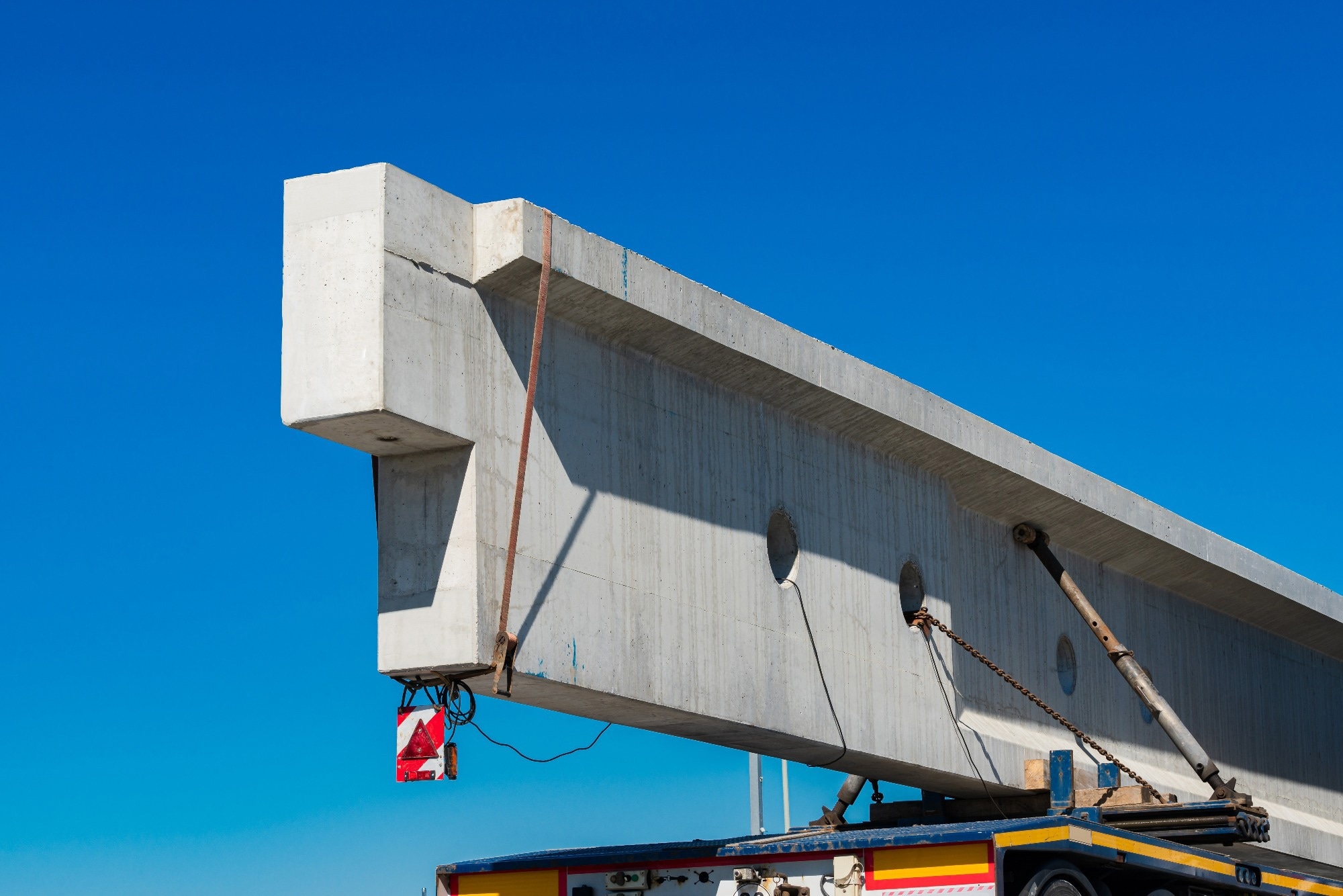 A large concrete beam is being lifted onto a flatbed truck during daylight. The scene highlights the scale of construction materials used in infrastructure development. Clear blue sky overhead.