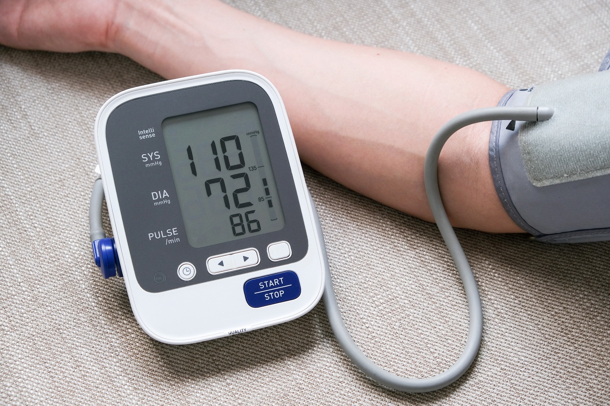 A man checks his blood pressure using a cuff device. The gray cuff is tight around his arm and the monitor reads SYS 110 mmHg and DIA 72 mmHg.